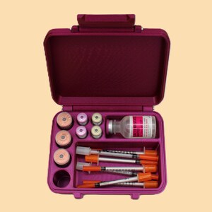 Puede incluir: Un estuche de viaje médico magenta abierto, que contiene viales, jeringas y una botella con una etiqueta roja. El estuche está organizado con compartimentos para cada artículo, lo que sugiere un kit de suministros médicos.