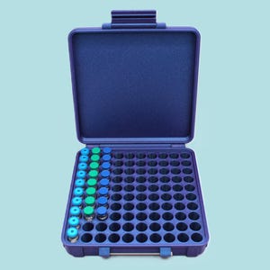 Op de afbeelding: Een donkerblauwe plastic opbergkoffer met een scharnierend deksel, voorzien van een raster met ronde openingen. Verschillende kleine glazen flesjes met felgroene en blauwe doppen zijn in de linkerzijde van het raster geplaatst, de rest is leeg.