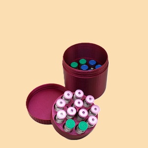 May include: A burgundy container with a matching lid, holding several small glass vials with pink and green caps. The open container reveals additional vials with blue and green caps. The vials are arranged in a circular pattern.