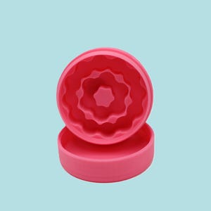 May include: A bright pink silicone donut mold with a flower-shaped center and wavy edges. The mold is in two parts, with the top piece showing the detailed interior design. The base is a simple, round shape.