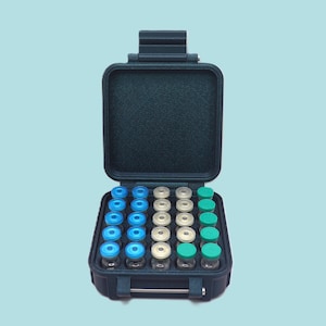 Op de afbeelding: Een open, teal-kleurige koffer gevuld met kleine glazen flesjes. De flesjes hebben blauwe, witte en groene doppen. De koffer is rechthoekig met een handvat en een sluiting. De achtergrond is lichtblauw.