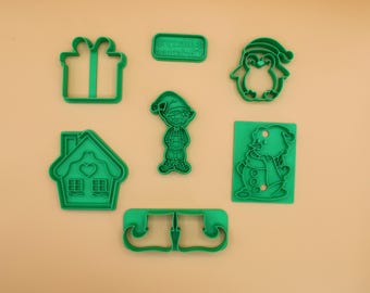 Puede incluir: Un conjunto de cortadores de galletas de plástico verde con formas navideñas, incluyendo un regalo, una casa, un elfo, un pingüino, un muñeco de nieve y un bigote. Los cortadores están sobre una superficie marrón claro.