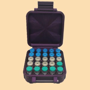 Op de afbeelding: Een paarse, open, harde koffer met een handvat, met meerdere kleine glazen flesjes met gekleurde doppen. De doppen zijn blauw, wit en groen. De koffer staat op een lichtbeige achtergrond.