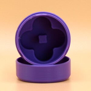 May include: A purple, 3D-printed cookie cutter with a four-leaf clover design. The cutter consists of a circular base and a top piece with the clover shape. The pieces are separated, ready for use. The background is a solid, light orange color.