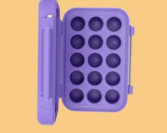 May include: A purple egg storage container, open to reveal 15 circular compartments. The container is rectangular with a hinged lid and a handle on the side. The background is a solid light orange color.