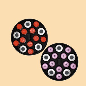 Puede incluir: Dos bandejas circulares negras llenas de pequeños viales. Una bandeja tiene viales rojos y plateados, la otra tiene viales rosas y plateados. Los viales están dispuestos en círculo.