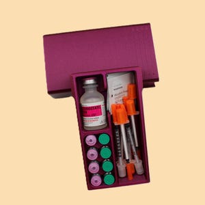 May include: A magenta medical kit containing a vial, syringes, alcohol prep pads, and several small vials. The kit is open, revealing the contents, and is set against a light yellow background.