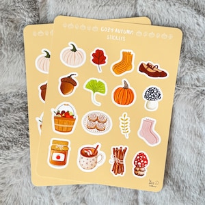 May include: Two sheets of stickers with a cosy autumn theme. The stickers feature pumpkins, leaves, socks, shoes, acorns, mushrooms, and other fall-themed illustrations. The text "COZY AUTUMN STICKERS" is printed at the top of the sheets.