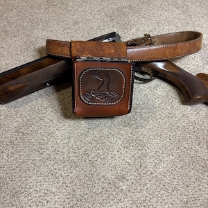 May include: A brown leather belt with a matching pouch featuring a duck design. The pouch is attached to the belt and is positioned over a shotgun. The belt and pouch are made of a distressed leather.