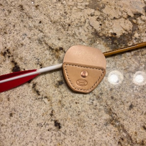 May include: A tan leather arrow rest with a stitched border and a copper rivet. A white arrow with red fletching is inserted into the rest. A wooden pencil is also visible.