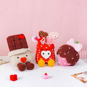 Puede incluir: Colecci&oacute;n de art&iacute;culos de ganchillo hechos a mano, que incluyen una barra de chocolate, una bolsa roja con caramelos en forma de coraz&oacute;n y una fresa. La barra de chocolate es marr&oacute;n y crema, la bolsa es roja con un coraz&oacute;n blanco y la fresa es rosa y marr&oacute;n.