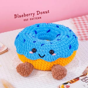 May include: A crocheted plush donut with a bright blue top, yellow bottom, and two brown feet. The donut has black safety eyes, a stitched smile, and pink cheeks. The text "Blueberry Donut PDF PATTERN" is visible above the plush object.