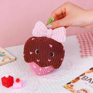 Puede incluir: Un cupcake de ganchillo hecho a mano con la parte superior marr&oacute;n, la base rosa y un lazo rosa. El cupcake tiene detalles de corazones y ojos negros. Est&aacute; sobre un mantel individual blanco.