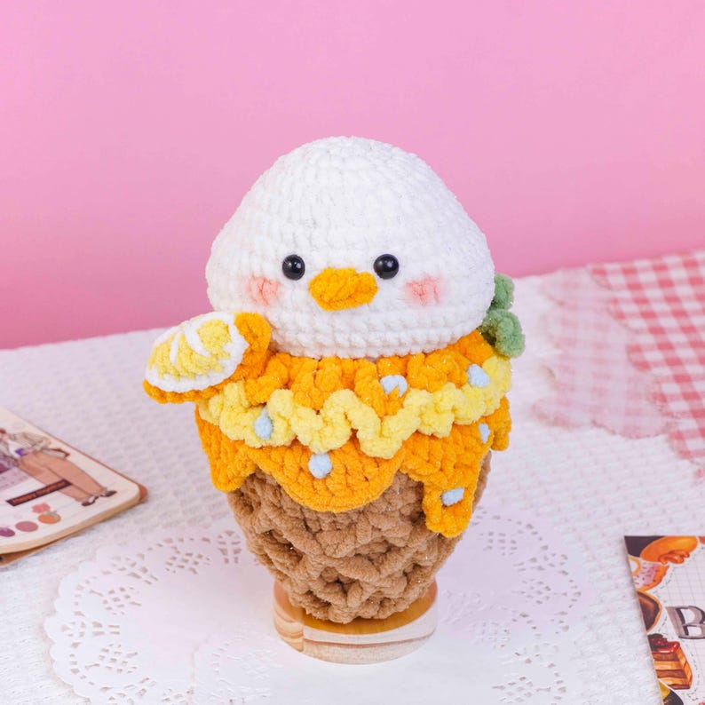 May include: A crocheted plush toy shaped like a dessert, featuring a white rounded head with black eyes, a yellow beak, and pink cheeks. The base is a textured brown cone topped with yellow yarn, a small lemon slice, and green accents.