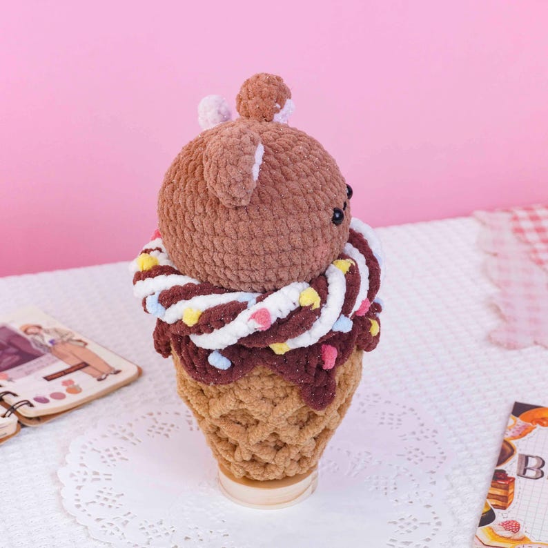 May include: A crocheted plush toy shaped like a bear head sitting atop a textured tan ice cream cone. The bear is brown with small ears, and a white and brown twisted yarn collar decorated with small, colorful pastel-colored yarn sprinkles.