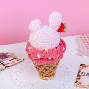May include: A crochet plush toy shaped like a strawberry ice cream cone. The top is light pink with two rounded ears and a small red strawberry accent. A thick, dark pink textured layer sits below, and the base is a tan waffle cone pattern.