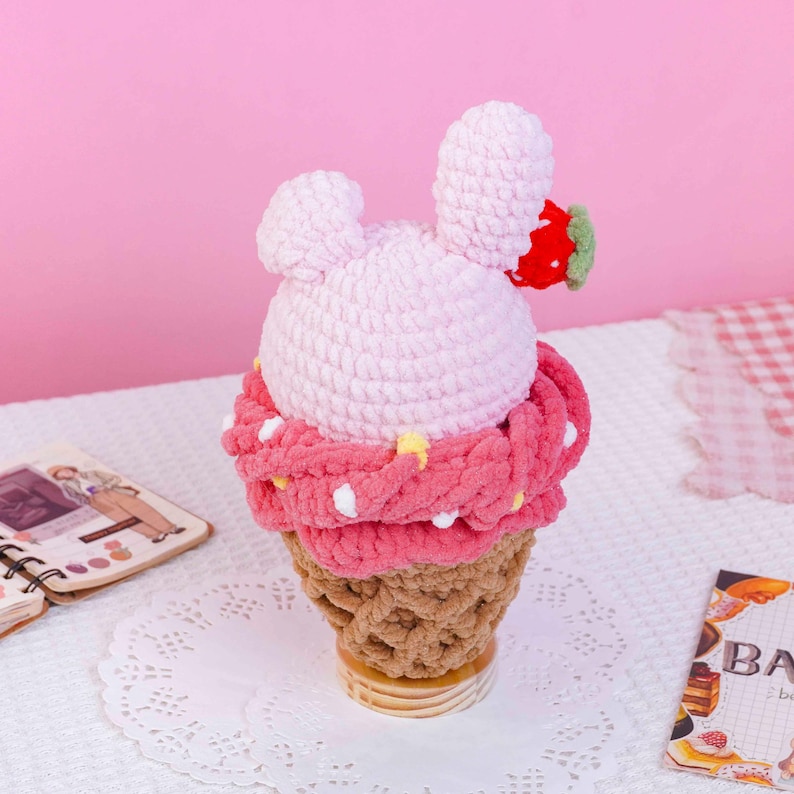 May include: A crochet plush toy shaped like a strawberry ice cream cone. The top is light pink with two rounded ears and a small red strawberry accent. A thick, dark pink textured layer sits below, and the base is a tan waffle cone pattern.