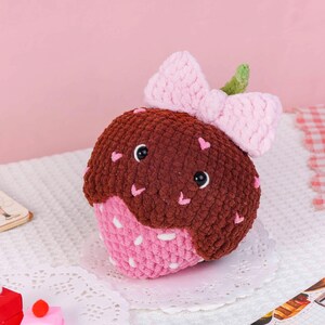 Puede incluir: Un cupcake de peluche hecho a mano, con la parte superior marr&oacute;n y la inferior rosa, decorado con peque&ntilde;os corazones rosas. Tiene un lazo rosa y un detalle de hoja verde. El cupcake tiene ojos negros y est&aacute; sobre un mantel blanco.