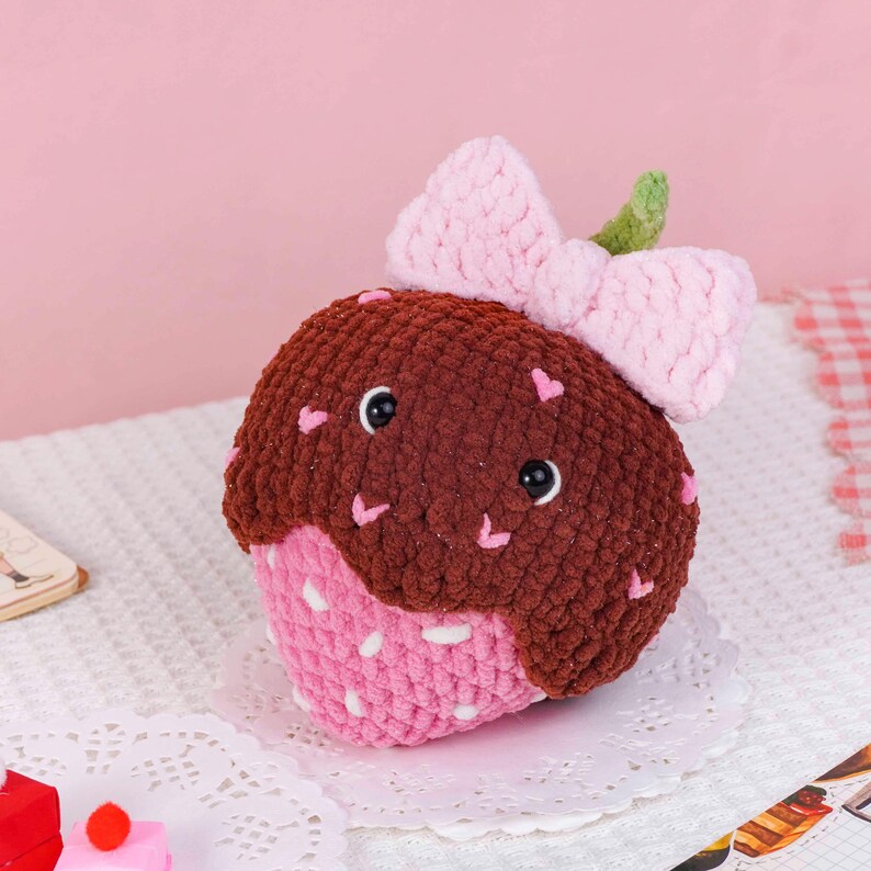 Puede incluir: Un cupcake de peluche hecho a mano, con la parte superior marr&oacute;n y la inferior rosa, decorado con peque&ntilde;os corazones rosas. Tiene un lazo rosa y un detalle de hoja verde. El cupcake tiene ojos negros y est&aacute; sobre un mantel blanco.