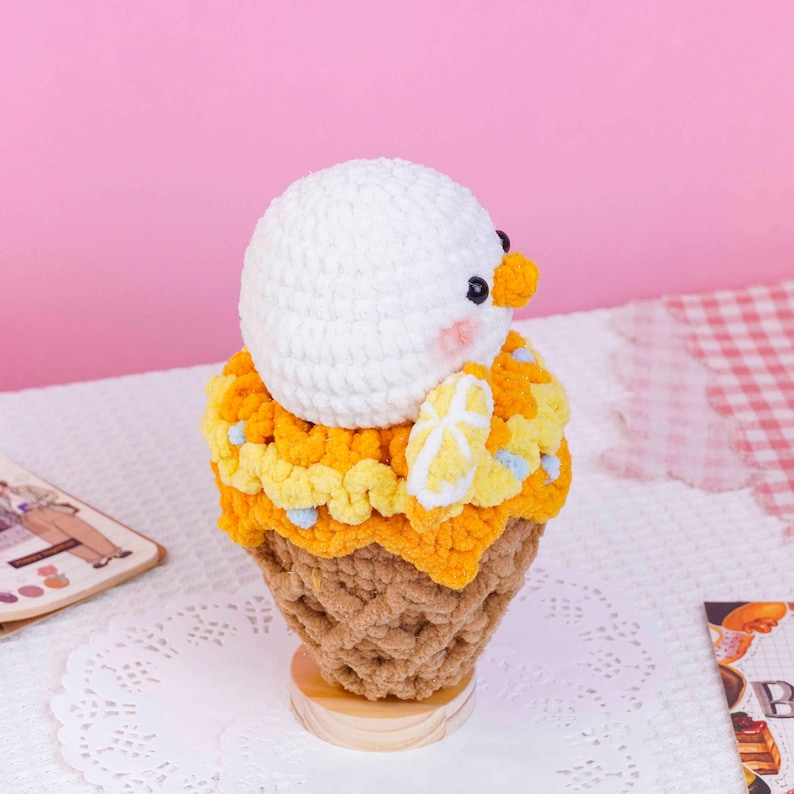 May include: A crocheted plush toy shaped like a scoop of white ice cream with a small orange beak and pink cheeks, topped with yellow textured yarn, sitting in a brown waffle cone base, all resting on a small wooden circular stand.