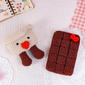 Puede incluir: Art&iacute;culos de ganchillo hechos a mano: una bolsa con forma de oveja de color crema con un coraz&oacute;n rojo y patas marrones, y una funda rectangular marr&oacute;n con un lazo rojo. Ambos art&iacute;culos est&aacute;n sobre una superficie blanca texturizada.