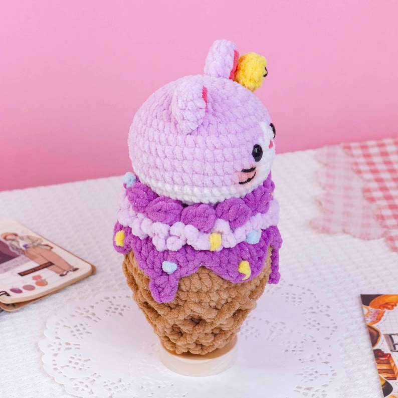 May include: A crocheted plush toy shaped like an ice cream cone with a light purple animal head on top. The head features small ears, a yellow accent, and embroidered facial details. The cone is tan, topped with dark purple textured yarn.