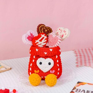Puede incluir: Una cesta roja hecha a mano con ganchillo, con una cara en forma de coraz&oacute;n blanco y pies de pomp&oacute;n amarillos. La cesta contiene varias piruletas en forma de coraz&oacute;n de ganchillo en varios colores y patrones, ideal para San Valent&iacute;n.