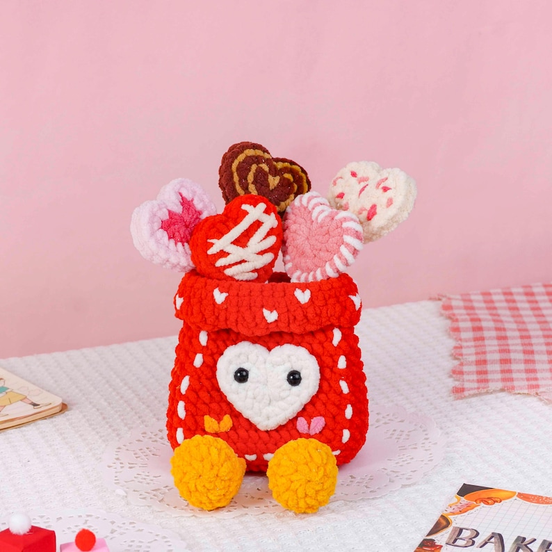Puede incluir: Una cesta roja hecha a mano con ganchillo, con una cara en forma de coraz&oacute;n blanco y pies de pomp&oacute;n amarillos. La cesta contiene varias piruletas en forma de coraz&oacute;n de ganchillo en varios colores y patrones, ideal para San Valent&iacute;n.