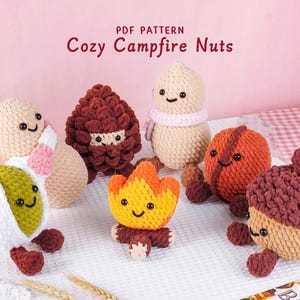 Op de afbeelding: Een verzameling handgemaakte gehaakte figuren, waaronder een dennenappel, een eikel en een kampvuur, met lachende gezichten. De figuren zijn in tinten bruin, oranje, geel en groen. De tekst "Cozy Campfire Nuts" staat bovenaan.