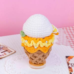 May include: A crochet ice cream cone toy featuring a textured tan waffle cone base, a bright yellow wavy layer, and a large, rounded white scoop on top. A small green accent is visible on the side of the white scoop, adding a touch of color to the soft, yarn-based construction of this handmade decorative item.