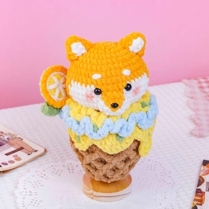 May include: A crocheted plush toy shaped like a fox head sitting atop a brown textured cone. The fox has orange and white yarn, black eyes, and pink cheeks. A yellow and light blue crocheted layer surrounds the base, with an orange slice accent.