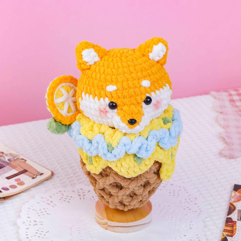 May include: A crocheted plush toy shaped like a fox head sitting atop a brown textured cone. The fox has orange and white yarn, black eyes, and pink cheeks. A yellow and light blue crocheted layer surrounds the base, with an orange slice accent.