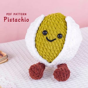 Op de afbeelding: Een handgemaakt gehaakt pistache-personage met een lachend gezicht, zwarte ogen en kleine rode voetjes. De pistache is groen en is omgeven door een witte schil. De tekst "PDF PATTERN Pistachio" is zichtbaar in de afbeelding.
