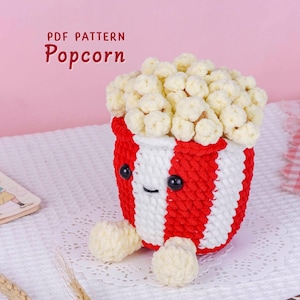 Może przedstawiać: Ręcznie robiona pluszowa zabawka amigurumi w kształcie popcornu z czerwono-białym paskowanym kubłem, wypełnionym puszystym, kremowym popcornem. Widoczny jest tekst "PDF PATTERN Popcorn". Pluszak ma czarne oczy i małe stopy.
