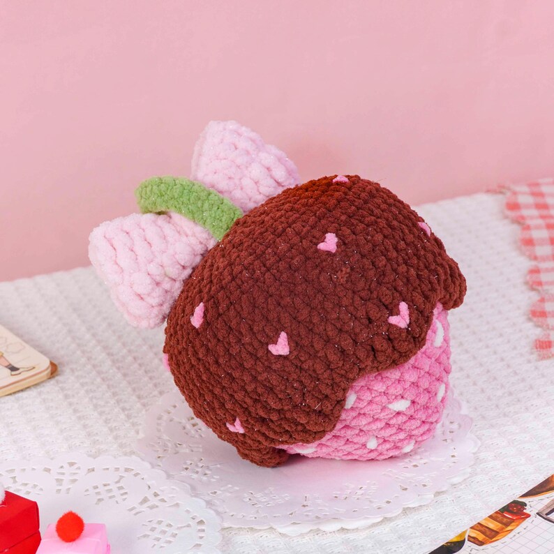 Puede incluir: Un juguete hecho a mano con forma de fresa de peluche. El juguete presenta una parte superior marr&oacute;n con detalles de coraz&oacute;n rosa, una base rosa y blanca con lunares y una hoja verde y un lazo rosa. El juguete est&aacute; hecho de hilo suave y texturizado.