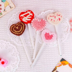 Puede incluir: Seis decoraciones hechas a mano en forma de coraz&oacute;n de ganchillo, en varios colores, incluyendo rojo, rosa y marr&oacute;n. Cada coraz&oacute;n est&aacute; unido a un palito blanco, similar a una piruleta. Los corazones est&aacute;n dispuestos sobre blondas blancas.