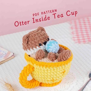 May include: A crocheted otter toy inside a yellow teacup. The otter is brown and white, holding a blue ball. The teacup has a yellow handle and rim. Text on the image reads "PDF PATTERN Otter Inside Tea Cup".