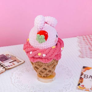 May include: A crochet plush toy shaped like a light pink bunny head sitting inside a tan waffle cone. The bunny features a red strawberry with a green leaf on its side. A thick, textured dark pink yarn layer surrounds the base of the bunny head.