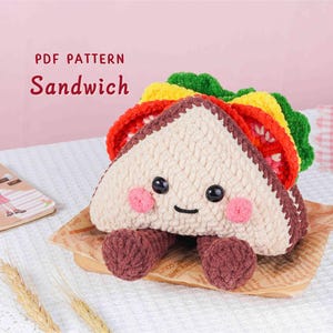 Puede incluir: Un sándwich de crochet con una cara sonriente, mejillas rosadas y pies marrones. El sándwich presenta capas verdes, amarillas y rojas, con el texto "PDF PATTERN Sandwich" en el fondo. Descansa sobre un trozo de papel marrón.