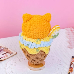 May include: A crocheted plush toy shaped like a scoop of orange ice cream in a brown waffle cone. The scoop is topped with a light blue ruffled trim, a yellow layer, and a small white and green slice of fruit attached to the side.