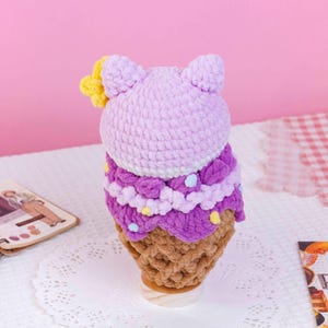May include: A crocheted plush toy shaped like an ice cream cone with a light purple rounded top, a darker purple ruffled middle section with small colorful dots, and a tan textured base. A small yellow flower is attached to the side.