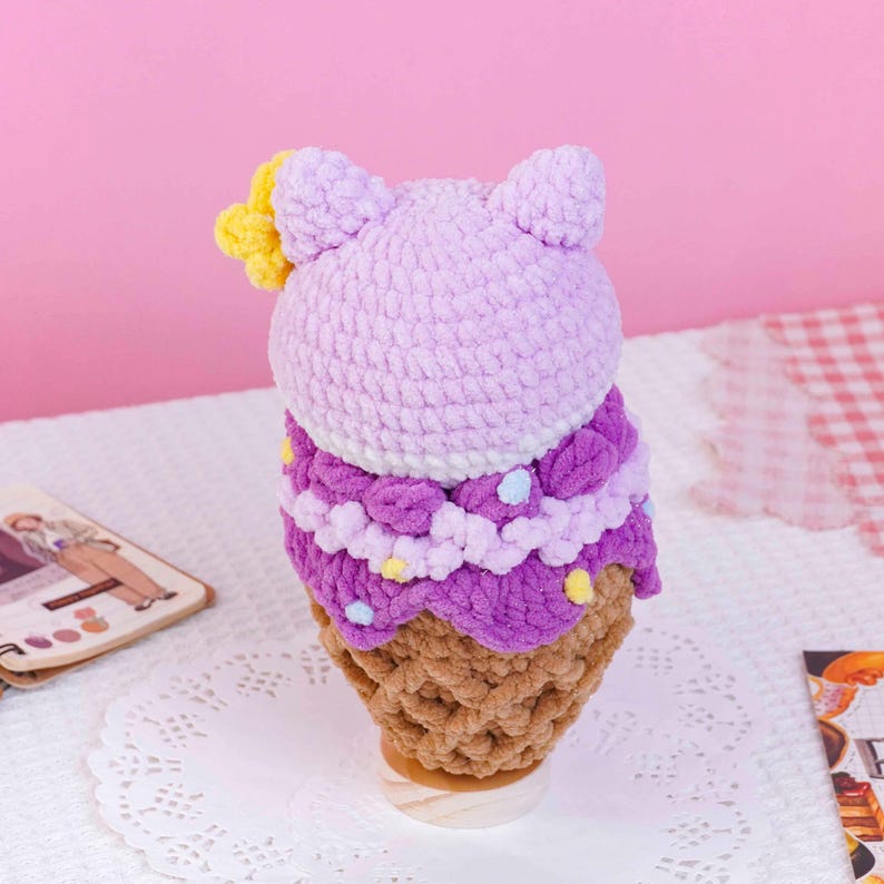 May include: A crocheted plush toy shaped like an ice cream cone with a light purple rounded top, a darker purple ruffled middle section with small colorful dots, and a tan textured base. A small yellow flower is attached to the side.