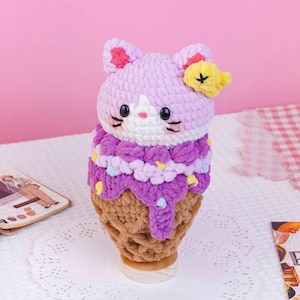 May include: A crochet plush toy shaped like a cat sitting in a waffle cone. The cat has light purple fur, a white face, black eyes, and a small yellow flower on its ear. The body is covered in dark purple yarn with small yellow and blue accents.