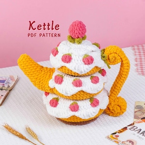 May include: A crocheted teapot in the shape of a cake, with yellow, white, and pink details. The teapot has a handle and spout, and the text "Kettle PDF Pattern" is visible.