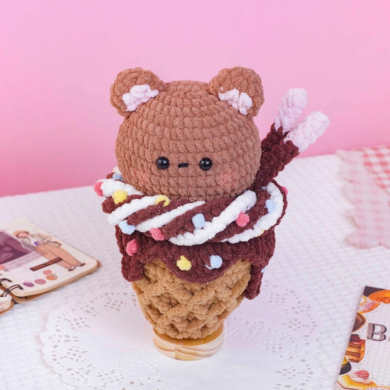 May include: A brown crocheted bear head sits atop a tan waffle cone base. The bear has small pink inner ears and black eyes. A dark brown crocheted layer with colorful pom-poms and a white twisted yarn accent wraps around the bear's neck.