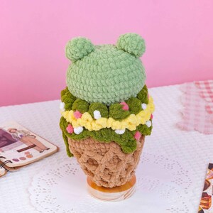 May include: A plush crochet ice cream cone toy featuring a light green rounded top with two small ears, a dark green middle section with yellow accents and small white and pink pom-poms, and a textured light brown cone base on a stand.