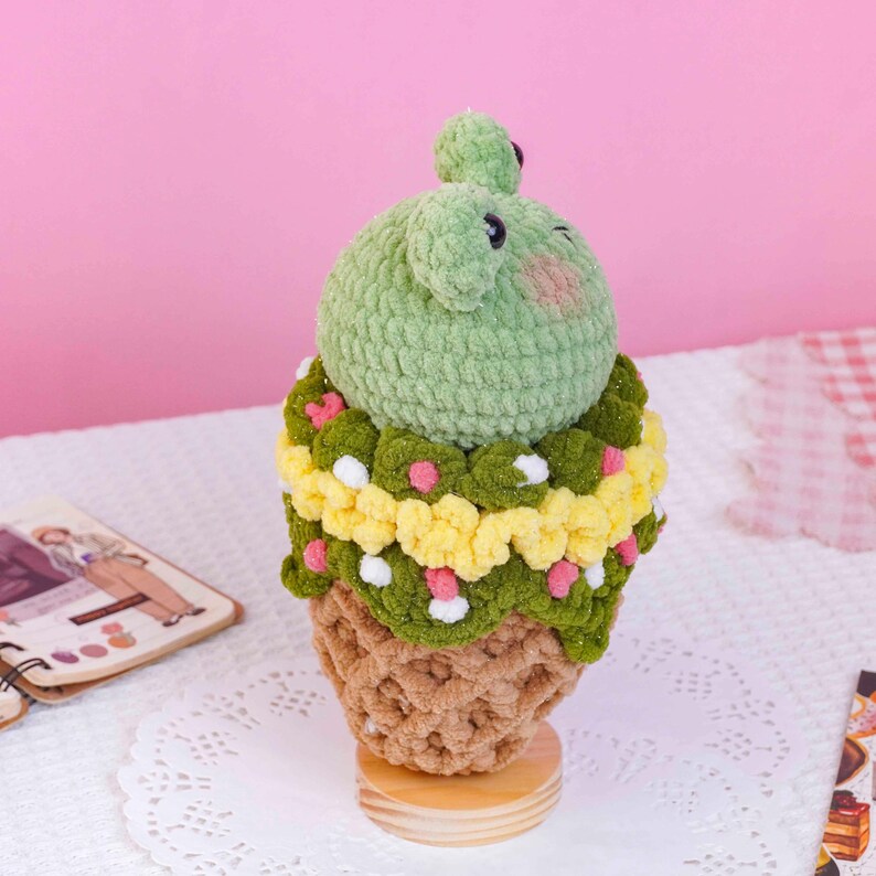 May include: A crocheted plush toy shaped like a frog sitting atop a dessert cone. The frog is light green with dark eyes and pink cheeks. The middle section features layers of dark green, yellow, and pink yarn, while the base is tan.