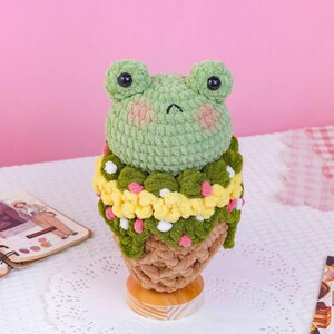 May include: A crochet plush toy shaped like a frog head sitting atop a tan textured cone. The frog is light green with black eyes and pink cheeks. A decorative collar of dark green, yellow, and white yarn loops surrounds the frog's neck.