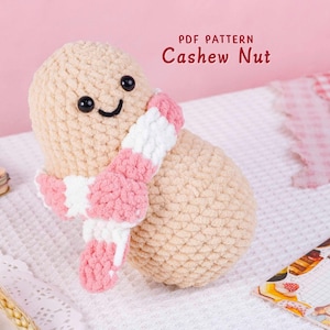 Puede incluir: Una muñeca de anacardo de ganchillo con una cara sonriente y una bufanda a rayas rosa, blanca y rosa. El texto "PDF PATTERN Cashew Nut" es visible en la imagen. La muñeca es beige y está sobre una superficie blanca.