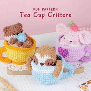 May include: Three crocheted tea cup critters: a bear in a yellow cup, a sloth in a blue cup, and a pink elephant in a purple cup. The text "PDF PATTERN Tea Cup Critters" is at the top. The critters are on a wooden board.